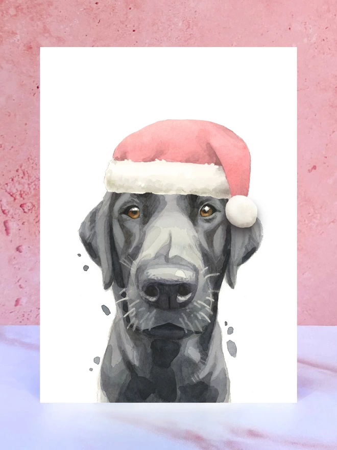 A Christmas card featuring a hand painted design of a black labrador, stood upright on a marble surface surrounded by pompoms. 