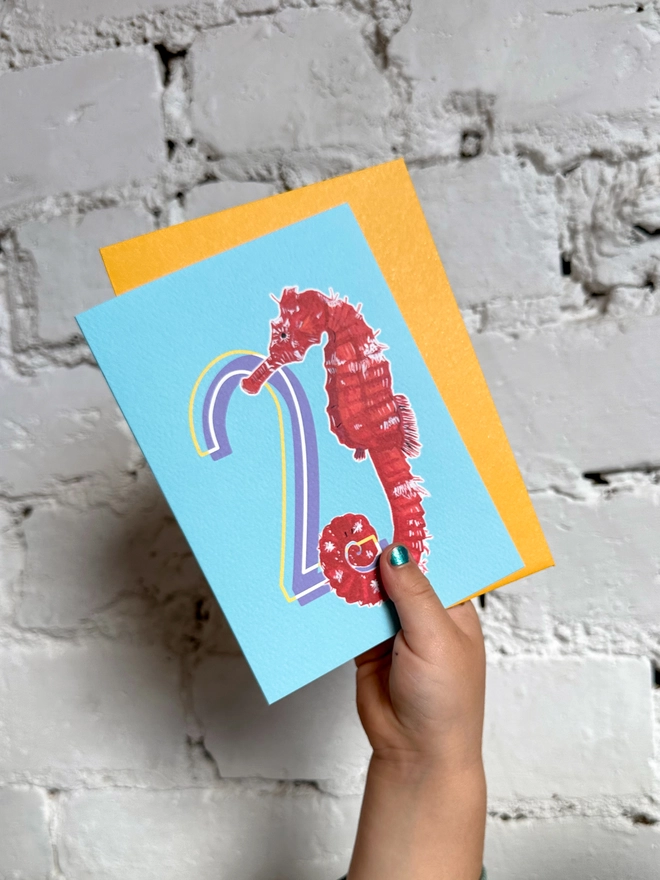 a bright blue card with a big purple number 2 and an illustration of a red seahorse and a bright yellow envelope