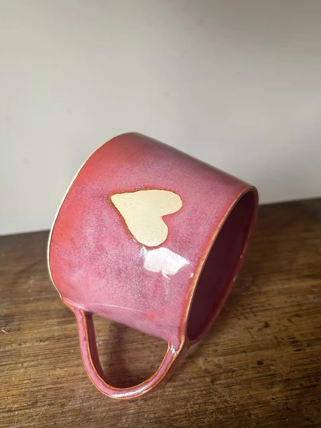 Heart Mug Raspberry Red, a red heart mug lying on its side on a wooden surface, in front of a plain background. 