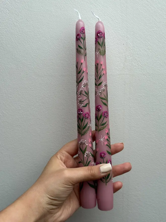 Pink Blooms Tapered Dinner Candle, a woman holding a pair of pink candles against a plain backdrop. The floral design wraps around the stalk. 