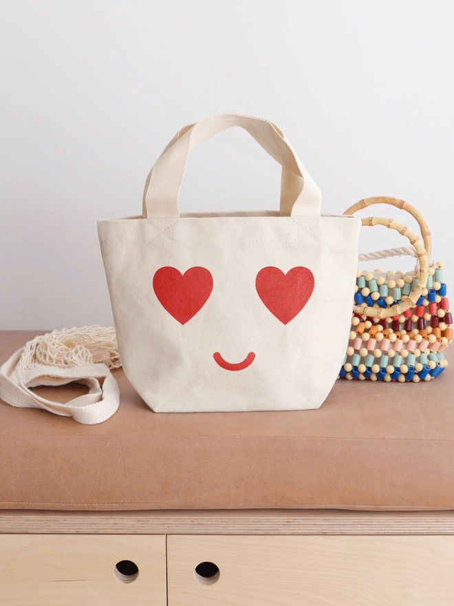 a mini kid's size tote bag with heart eyes and smiling mouth sitting on a bench