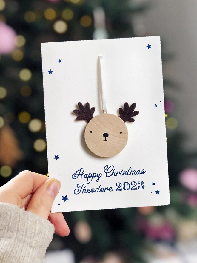 A personalised Christmas greetings card with a small wooden reindeer decoration attached is being held in front of a Christmas tree.