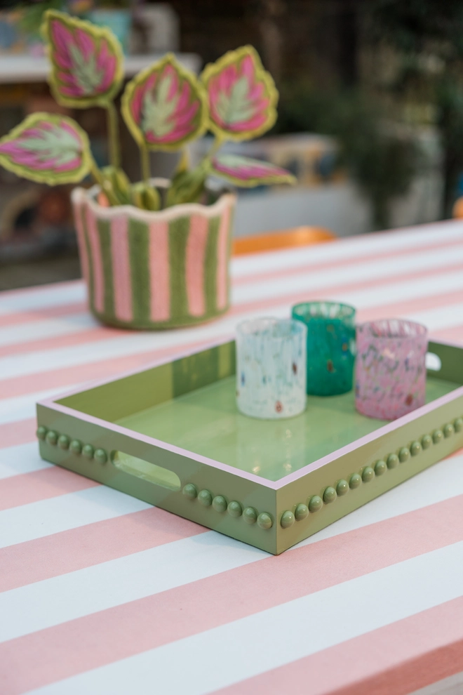 Hand Painted Green Bobbin Tray with Pink Edge and Handles on a pink and white striped tablecloth, with a felt plant in the background. There are 3 colourful glasses sat on the tray