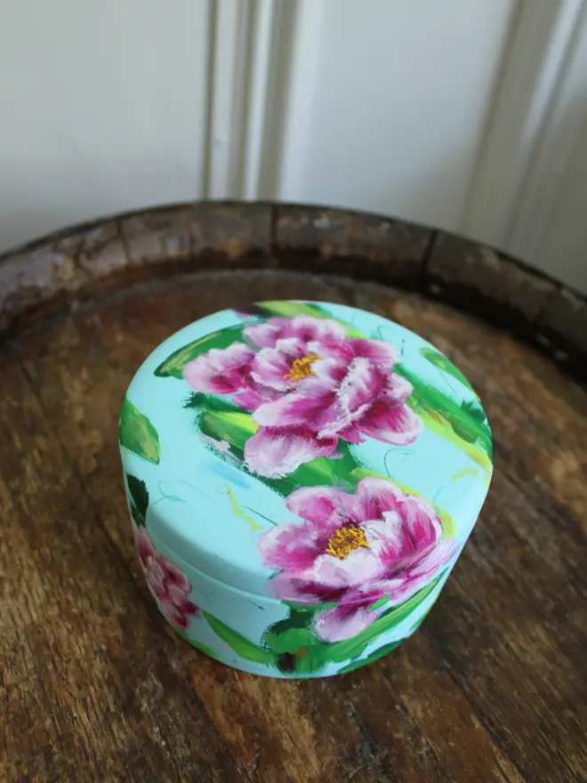 A beautifully hand painted floral ceramic round box background painted in a pale blue adorned in vibrant pink and white peony flowers.
