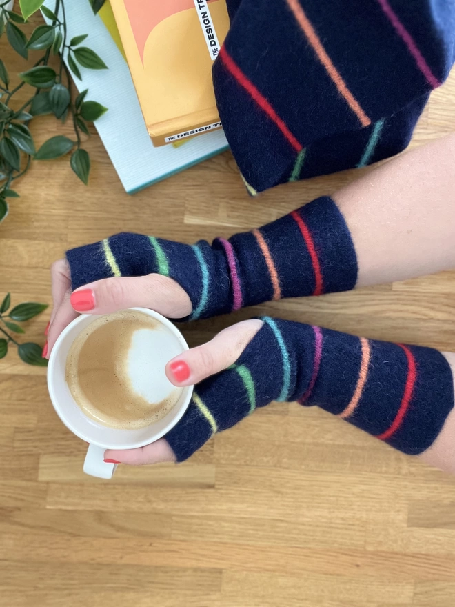 Knitted navy wristwarmers with multicoloured thin stripes, worn and shown with hands holding a coffee