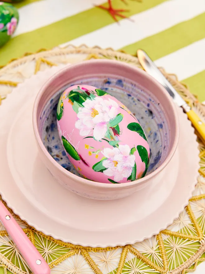 Soft blue hand painted wooden egg as part of a wider tablescape, positioned in a blue speckled bow on a blue scalloped plate. These are resting on a green jute tablemat atop a green and white striped tablecloth