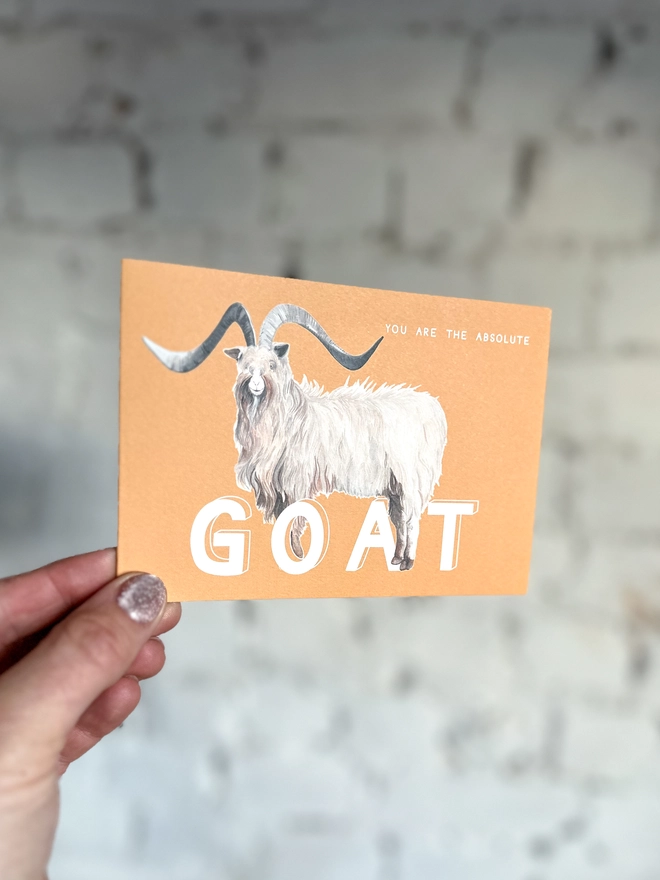 an orangey yellow greetings card featuring an illustration of a bilberry goat with the text “you are the absolute GOAT”