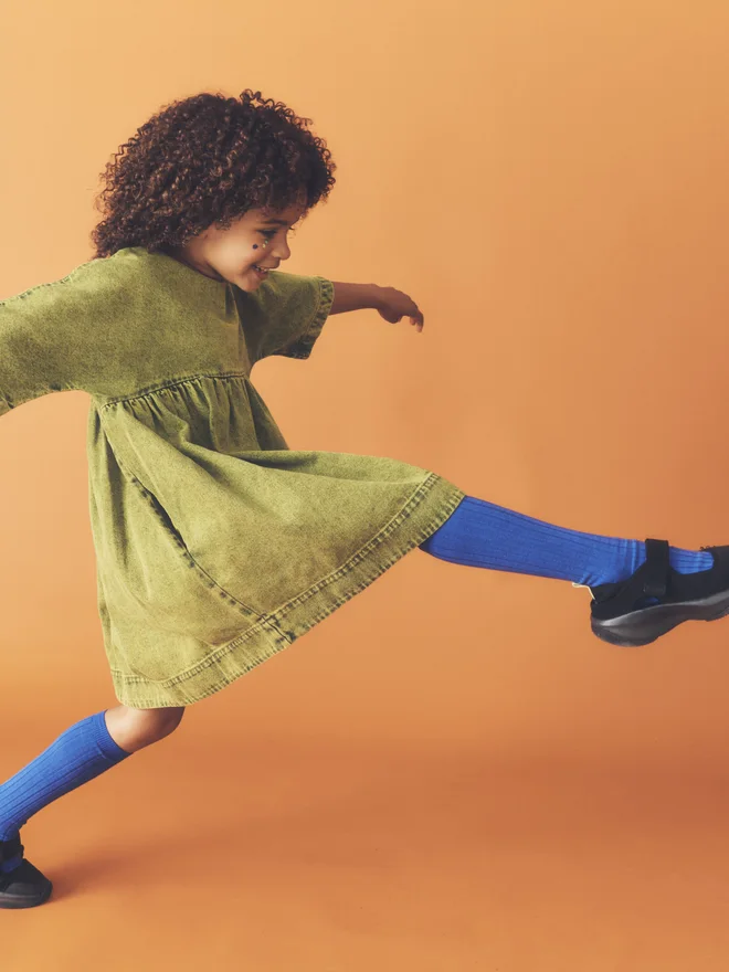 Side view of a child mid-leap against a burnt orange backdrop, wearing the Studio Koter Rumi dress in green acid-wash denim with cobalt knee socks.