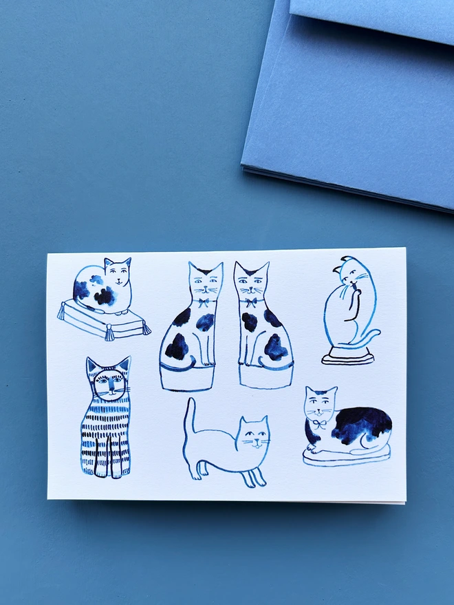 An illustrated greeting card featuring ink drawings of ceramic cats with a pale blue envelope