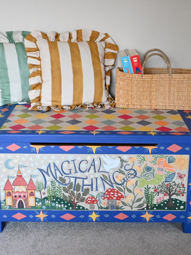 A hand-painted blue chest, likely a toy chest or storage chest, featuring a colorful diamond pattern on the top and a whimsical scene including a castle, trees, mushrooms, and other fairytale-inspired imagery on the front. The phrase "Magical Things" is written in a playful font. Two striped pillows, one green and one gold, and a natural woven basket with two children's books inside rest on the closed lid. The chest is centred against a neutral background.