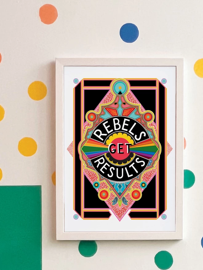 Rebels Get Results is written in white on a black background at the centre of this vibrant, abstract portrait illustration, with a black background and rainbows emitting from the centre, and multi-coloured detailing. The picture is hanging in a white frame on a white wall, with yellow, orange, green and blue spots and a green and yellow rectangle painted in the top left hand corner.
