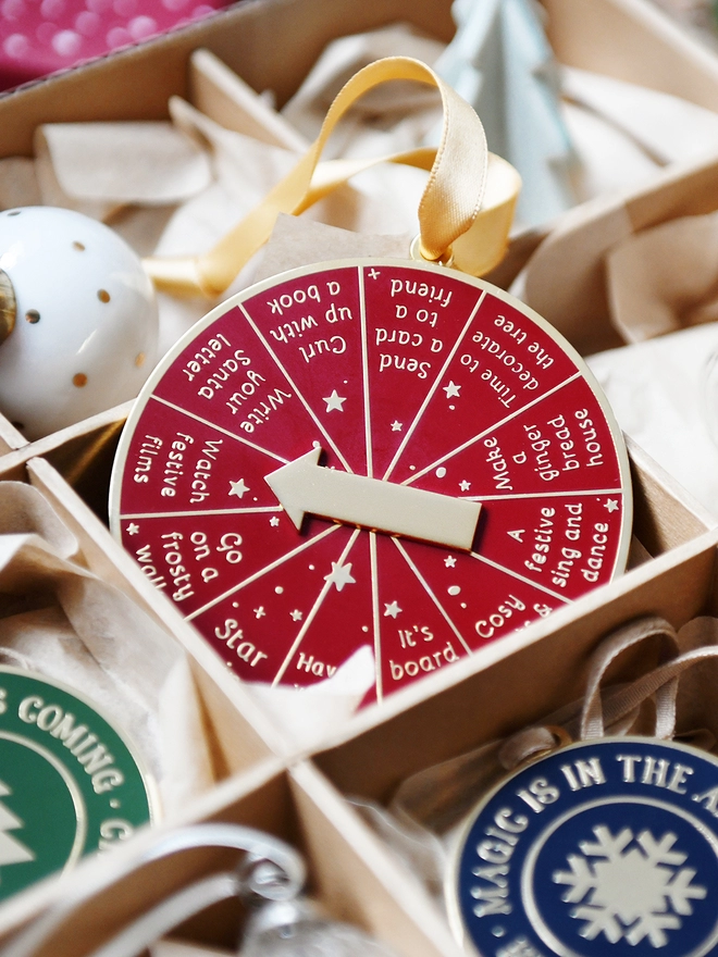 A deep red and gold Christmas decoration is tucked into a decorations box. It has 12 segments, each one with a different Christmas activity idea, and a golden arrow in the centre.