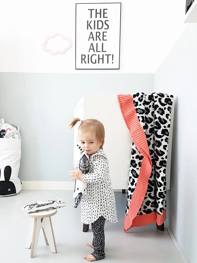 A bright and bold modern nursery with a toddler standing in the foreground in front of a cot. Over the cot is draped a black and white leopard print blanket with coral pink trim. In the background is a black and white typography poster that reads The Kids Are All Right!.