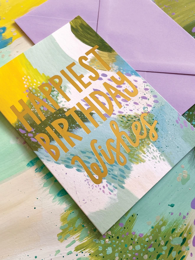 The colourful Raspberry Blossom birthday card sits on top of a lilac envelope and the painting used to create its vibrant design