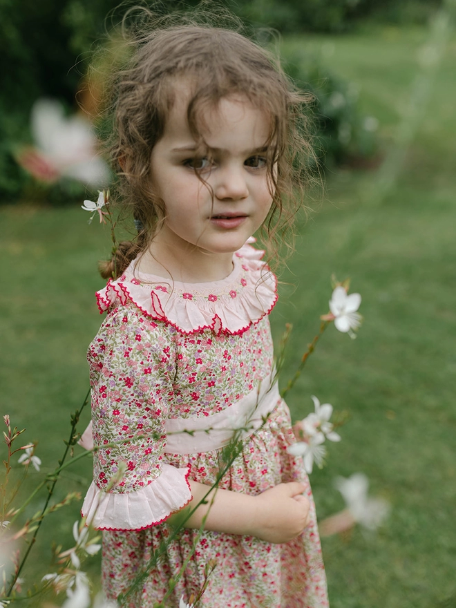 A little girl is surrounded by flowers wearing a floral dress with a pink frill collar and hand embroidered detailing