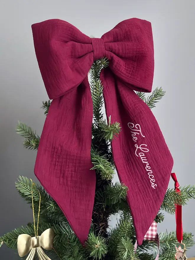 A handmade burgundy Christmas bow tree topper sits at the top of a decorated Christmas tree.