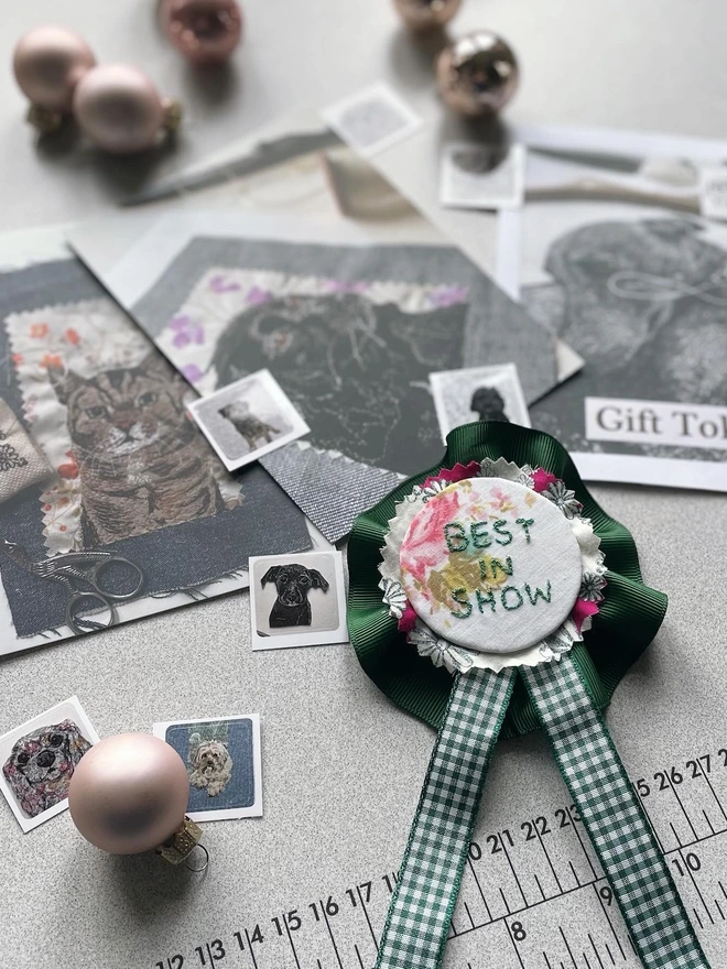 a handmade rosette, with "best in Show" embroidered on it, some stickers and postcards in the background