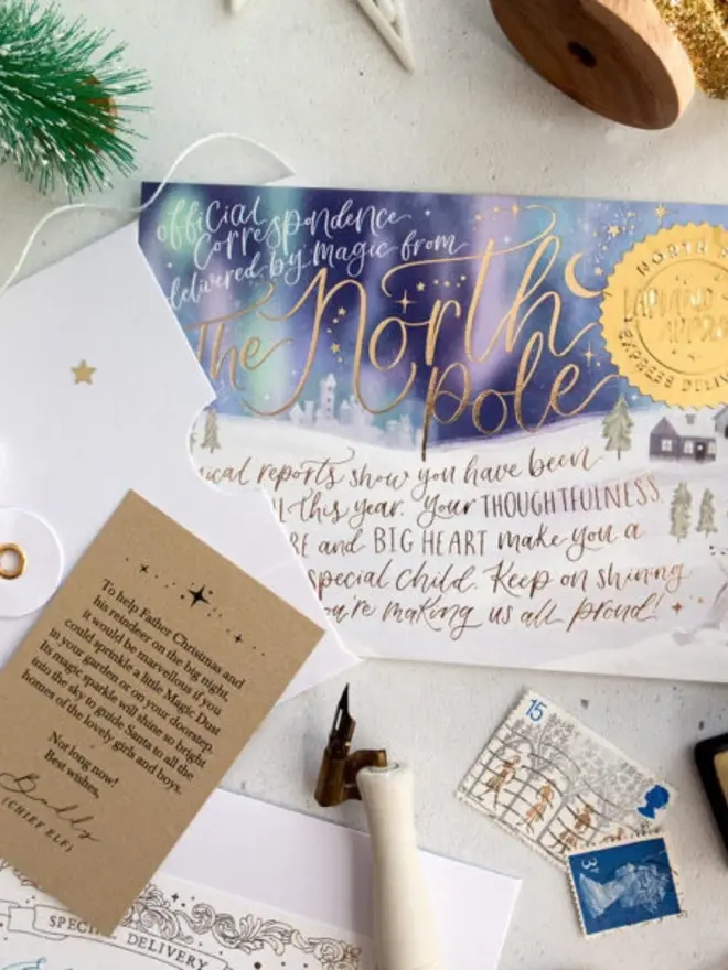 A close-up of the North Pole notecard with gold foil hand lettering reading "Magical reports show you have been wonderful this year. Your thoughtfulness, kind nature and big heart make you a very special child", with a kraft instruction card, a dip pen and vintage stamps.