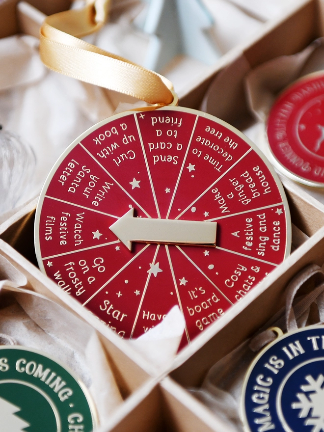 A deep red and gold Christmas decoration is tucked into a decorations box. It has 12 segments, each one with a different Christmas activity idea, and a golden arrow in the centre.