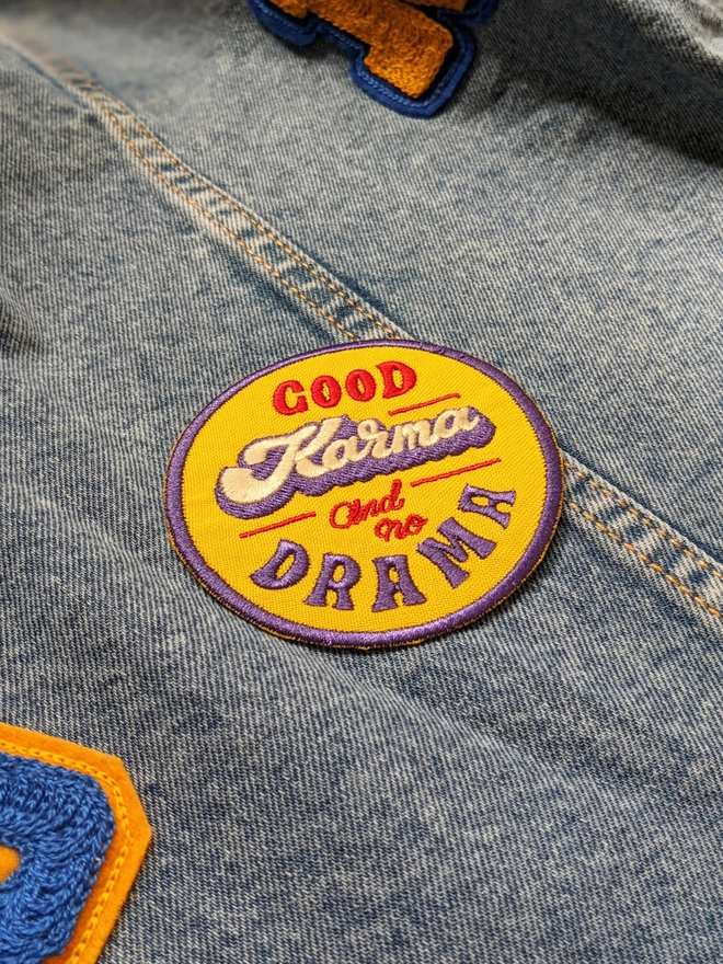 Yellow, Blue & Red round typographic patch reading 'Good Karma and No Drama'. The word Karma is written in a thick cursive lettering style in white, with a blue drop shadow, while the additional type is written in a retro style capital lettering & a smaller red cursive making up a fun retro style fabric patch. 