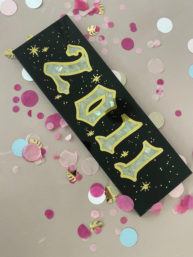 Personalised Year Date Sign, a black and gold personalised year sign sitting on a plain surface joined by confetti. 