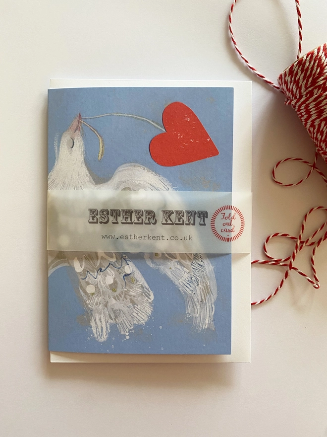 Esther Kent greetings card showing white flying dove in a light blue background, carrying a vibrant red heart on a string