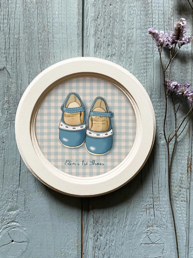 a photograph of a framed personalised illustration of a pair of blue shiny baby shoes, 1st shoes framed in a beautiful ivory painted wooden frame 