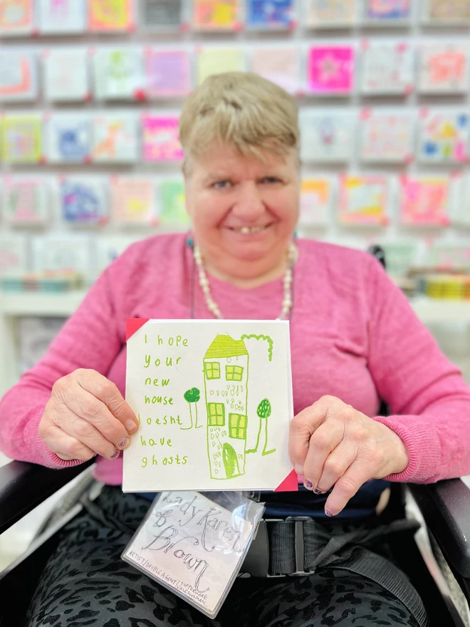 Artist holding I Hope Your New House Doesn't Have Ghosts card with a green house & garden design
