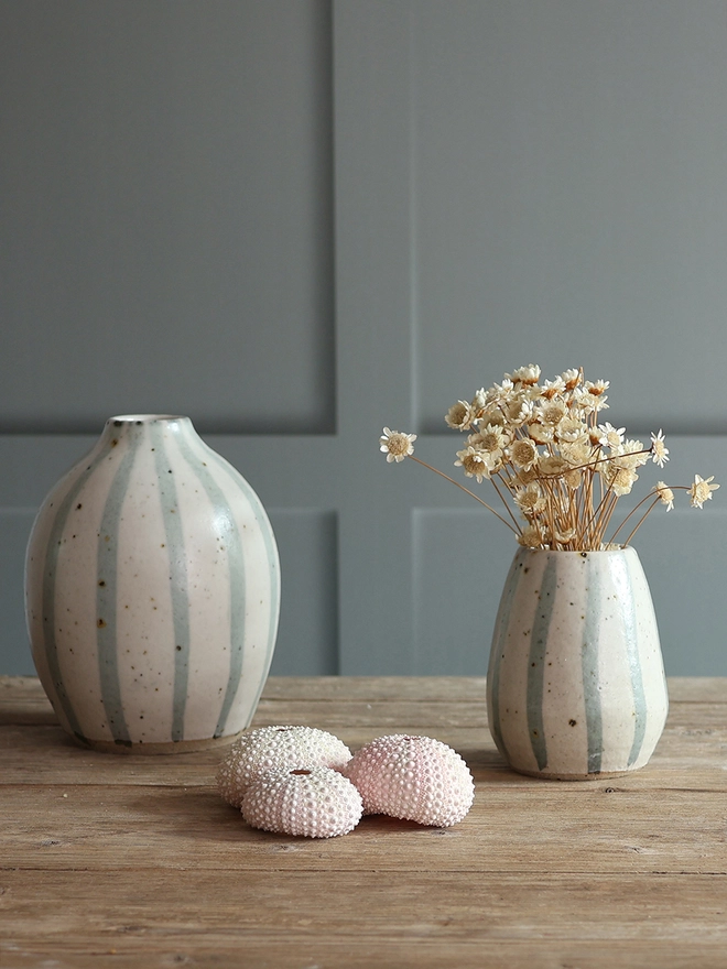 Handmade Ceramic Stoneware Small Vase in Blue and White Stripe filled with dried flowers sat on a wooden surface alongside our Handmade Stoneware Ceramic Rounded Vase in Blue and White Stripe. A collection of three sea urchins sit in-between the vases. 