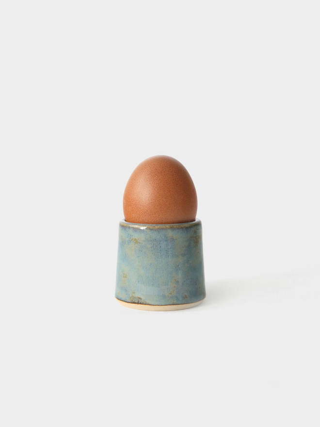 A handmade ceramic egg cup in seafoam blue glaze holding a brown egg, photographed against a plain white background
