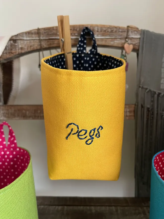Yellow storage bag hanging on a wooden stand, the front embroidered with ‘Pegs’ in navy thread.