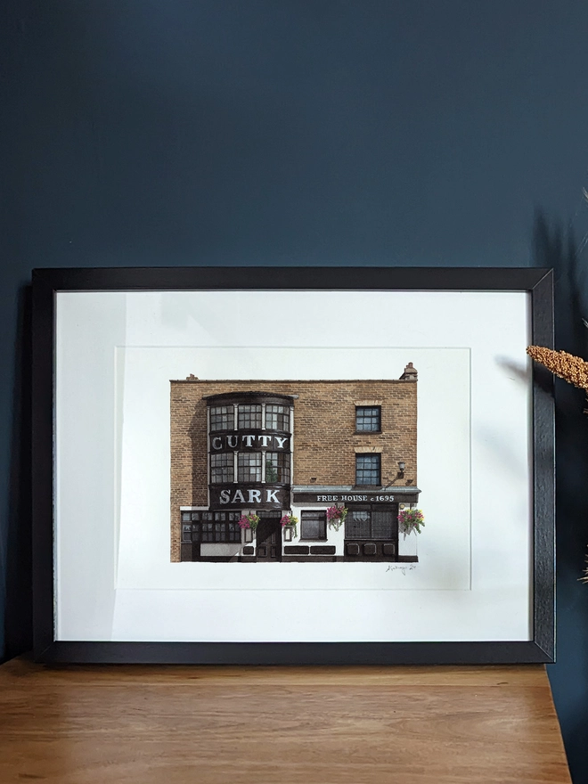 A watercolor print of the Cutty Sark pub displayed on a white mantelpiece next to a dark blue glass vase.