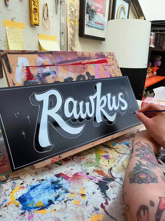 Hand-painting of an almost finished sign spelling out 'Rawkus' in white on black.