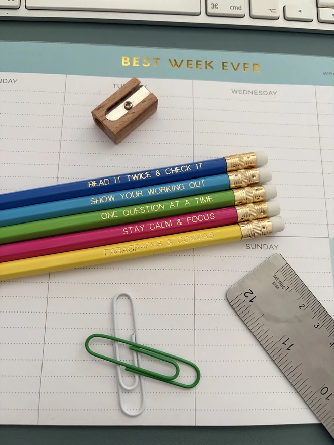 Exam essentials pencil set - Five colourful motivational exam pencils with gold foil lettering are styled on a pastel desk setup with stationery and a weekly planner.
