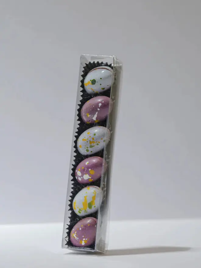 Clear rectangular box filled with assorted solid milk and dark chocolate mini eggs in pastel colours with speckled paint-style decoration, displayed on a white background.