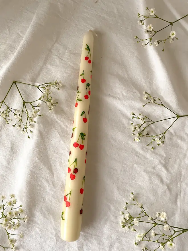 Mon Cherry & Tulip Fever Candle, a white candle lying on a table cloth accompanied by flowers. It's floral design wraps around the stalk. 