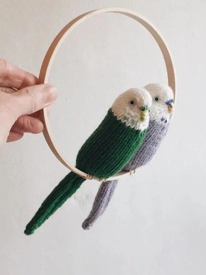 Colourful Hand Knitted Budgerigar, a duo of colourful birds sitting in a wooden ring against a white backdrop. 