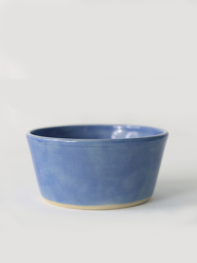 A single hand-thrown ceramic breakfast bowl in pacific blue, with a glossy glaze showing subtle variation in tone, a gently flared form and unglazed natural clay base, photographed against a plain light grey background.