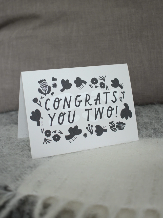 Black and white greeting card with illustration and the words Congrats you two written on it stood up with grey cushions and a blanket 