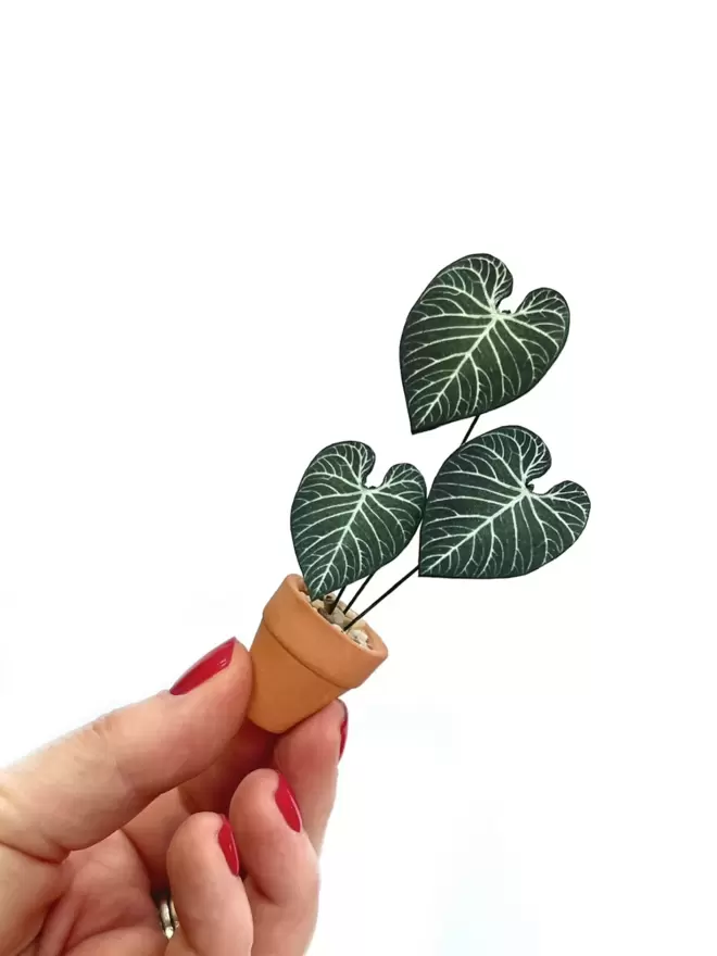 A miniature replica Anthurium Regale paper plant ornament in a terracotta pot held between 2 fingers against a white background