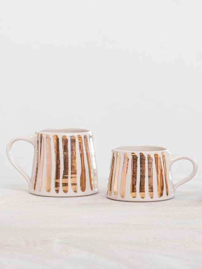 Glossy handmade mug with hand painted gold stripes on a blurred white background, both tall and short versions