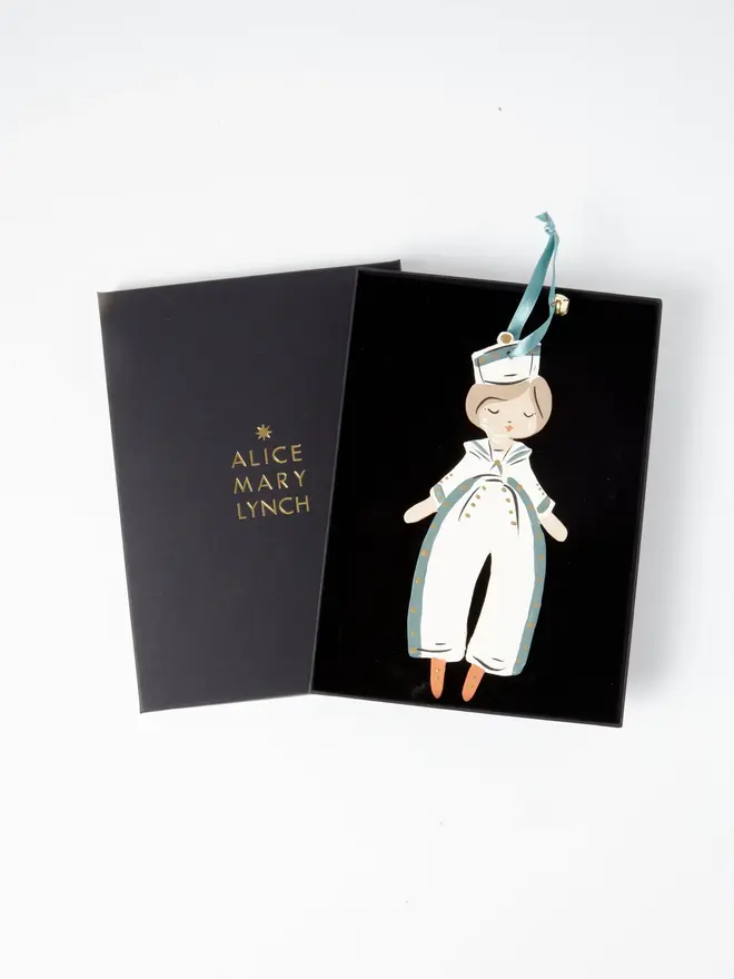 Sailor Heart Tree Dancer. wooden hanging sailor boy decoration in an open black gift box embossed "alice mary lynch" in gold, with a white sailor hat, cream and duck egg blue jumpsuit with gold buttons and orange shoes