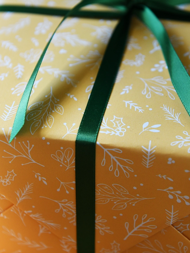 A gift wrapped in yellow wrapping paper with a gentle botanical design has a green ribbon tied around it.