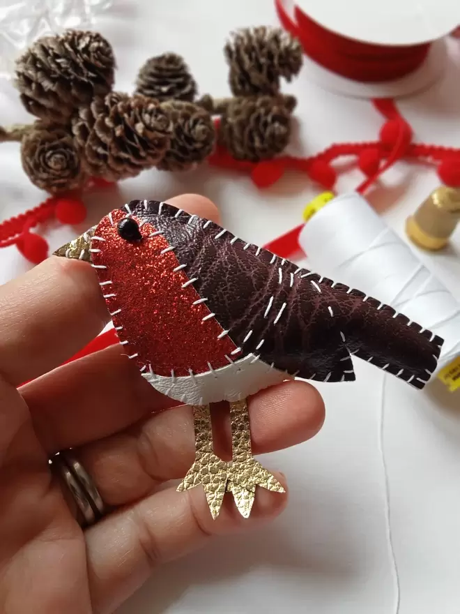 A hand holding a leatherette robin brooch, hand stitched, with red glittery breast, sits amongst pine cones, red ribbon, pom pom trim, thimble and white reel of cotton