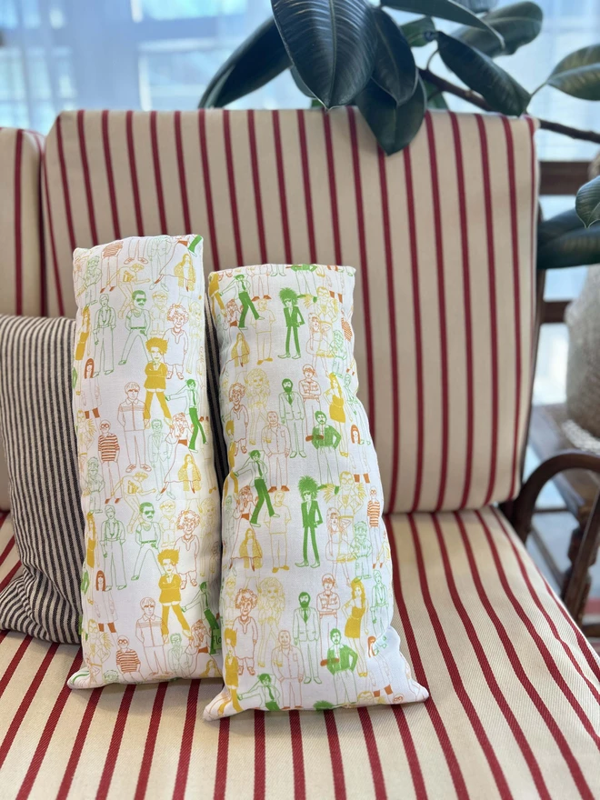 The back of two cushions featuring a design of celebrities in yellow, green and orange on white fabric.  The cushions are on a striped sofa surrounded by plants.