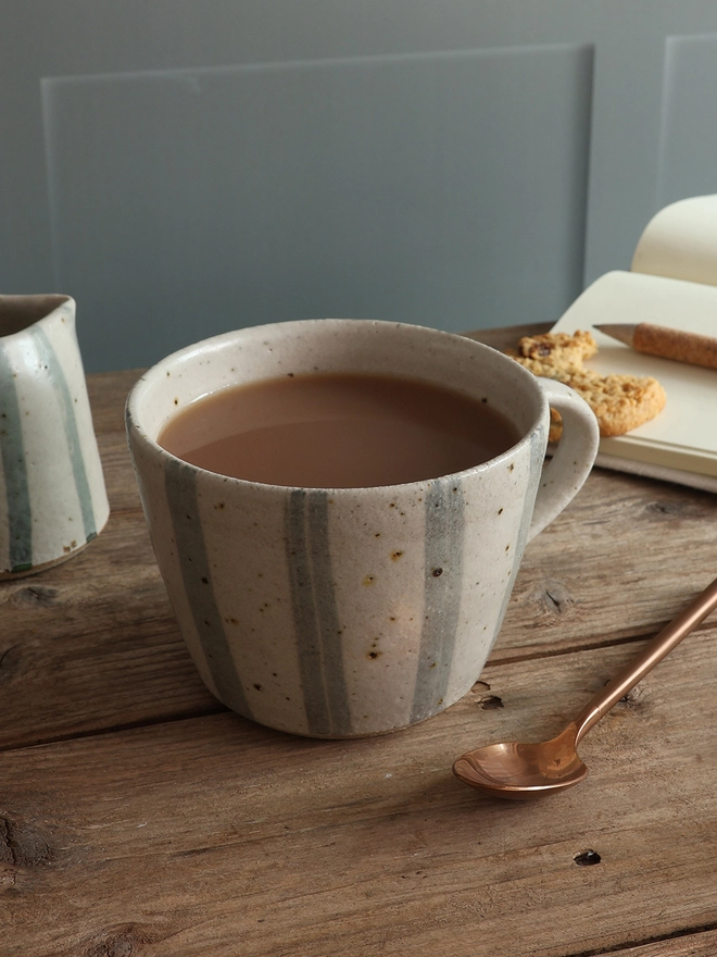 Handmade Ceramic Stoneware Mug in Blue and White Stripe sat on a wooden surface alongside a small handmade jug in the same style, a brass spoon, as note book with a pen and cookies sat on top. 