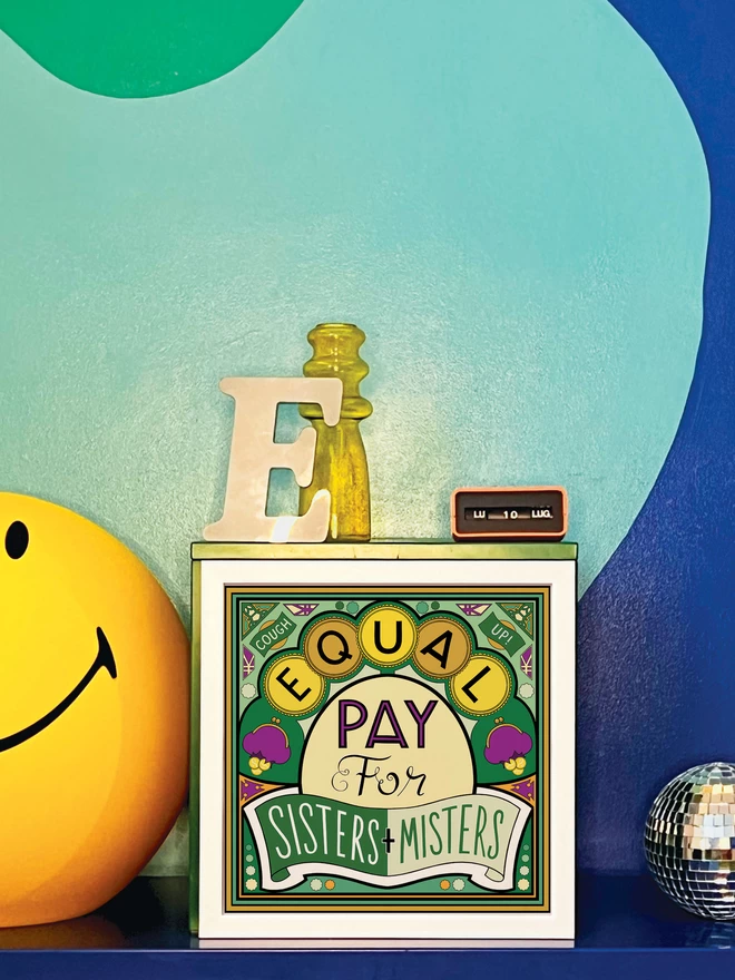 Equal Pay for Sisters and Misters is written over this green and yellow illustration. In the corners “Cough” and  “Up!” appears in banners at the top corners. The print is in a white square frame and is propped against a turquoise and dark blue wall. Next to the frame is a disco ball, a letter ‘E’ ornament, a yellow glass vase, an orange Italian plastic calendar showing the date as ‘LU 10 LUG’ and a large light up yellow Smiley lamp.