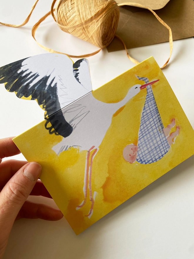 A new baby card by Esther Kent showing a black and white stork carrying a baby in a blue checked cloth, on a yellow background