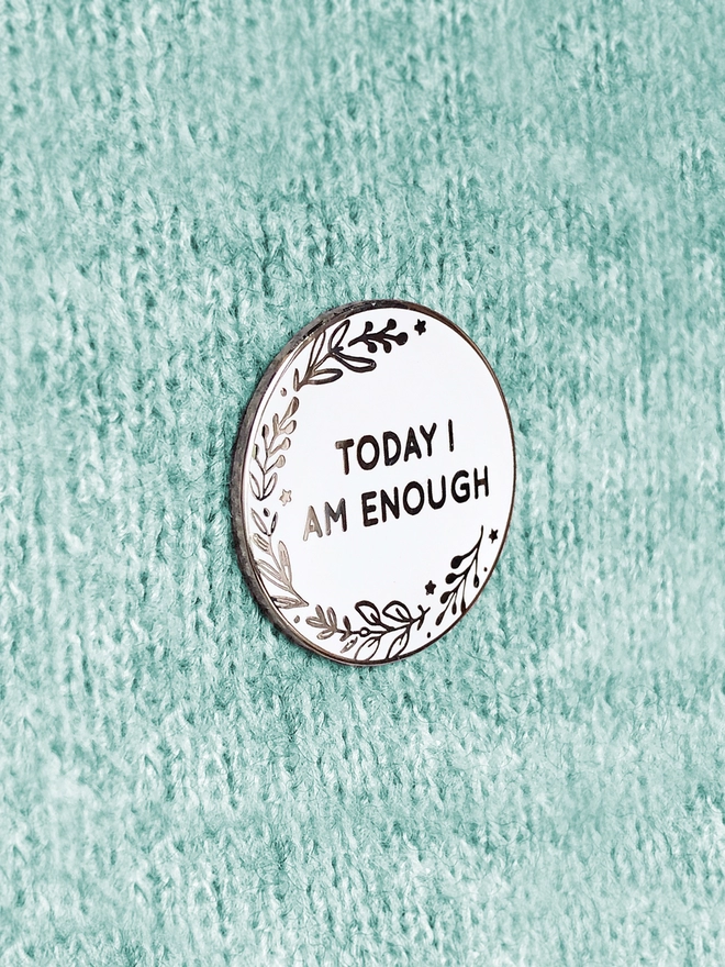 A round white enamel pin with a botanical design and the words "Today I Am Enough" is pinned to green fabric.