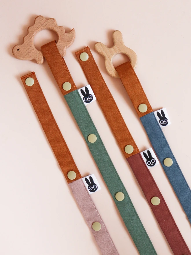 Handmade toy straps in woodland shades, featuring sturdy snap fastenings and small fabric tags with a bunny logo, neatly arranged in a row.